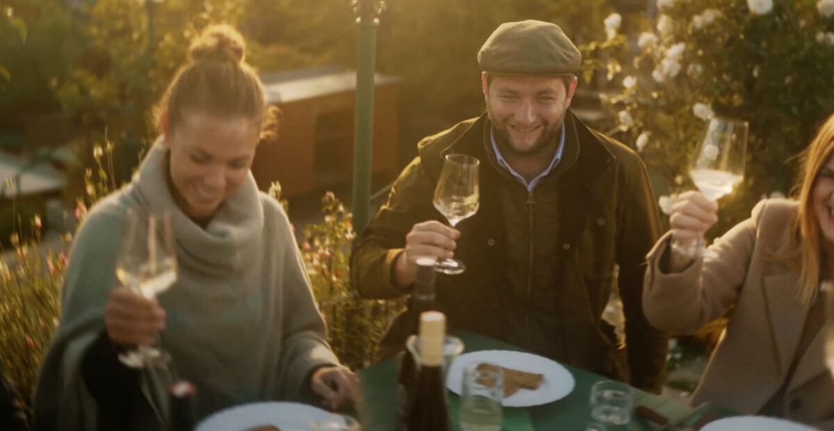 Drei Personen sitzen im Freien, lächeln und stoßen mit Weingläsern an einem Tisch mit Essen an.