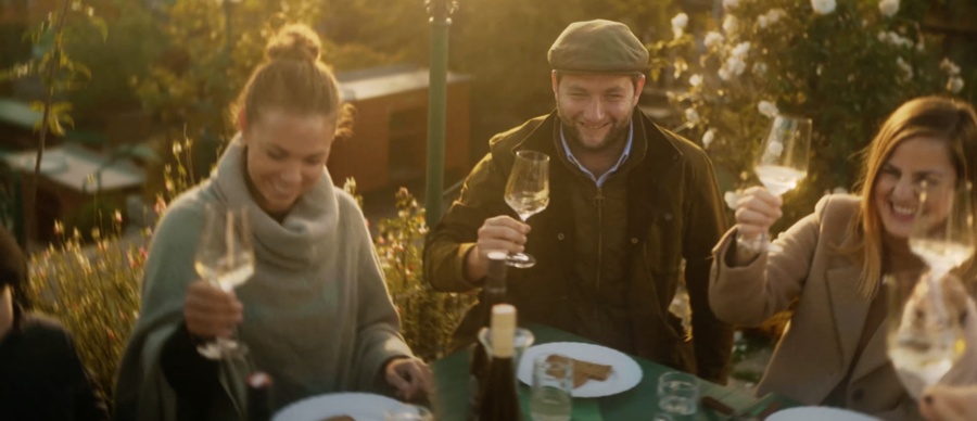 Drei Personen sitzen im Freien, lächeln und stoßen mit Weingläsern an einem Tisch mit Essen an.