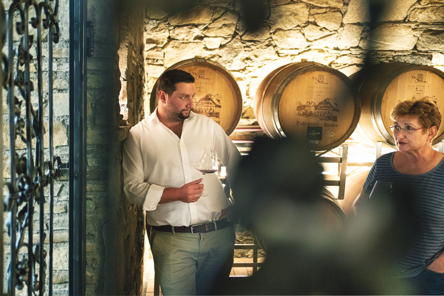 Ein Mann und eine Frau unterhalten sich in einem Weinkeller mit Gläsern in der Hand, hinter ihnen sind Fässer gestapelt.