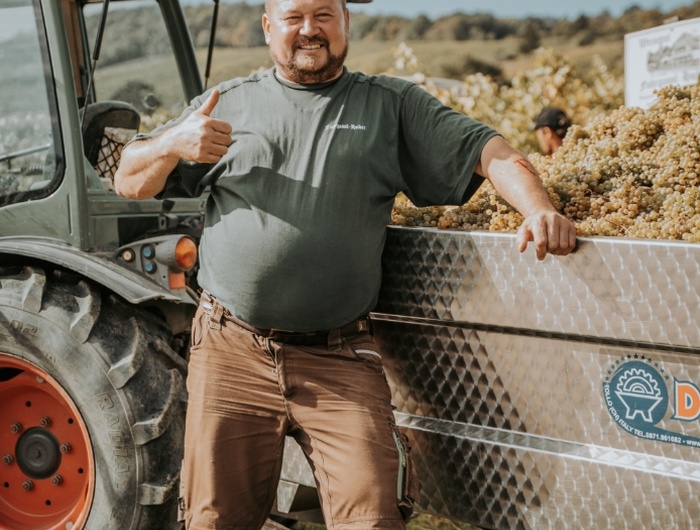 Ein Mann, der an einem sonnigen Tag neben einem Traktor und einem mit Weintrauben gefüllten Anhänger steht, lächelt und zeigt die Daumen nach oben.