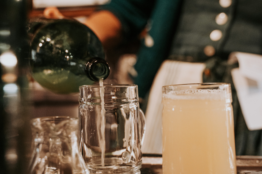 Eine Person gießt ein helles Getränk aus einer Flasche in einen Glaskrug, der neben einem gefüllten Becher steht.