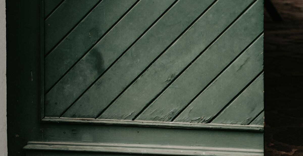 Nahaufnahme einer grünen Holztür mit diagonalen Paneelen und sichtbaren Anzeichen von Abnutzung und abblätternder Farbe.