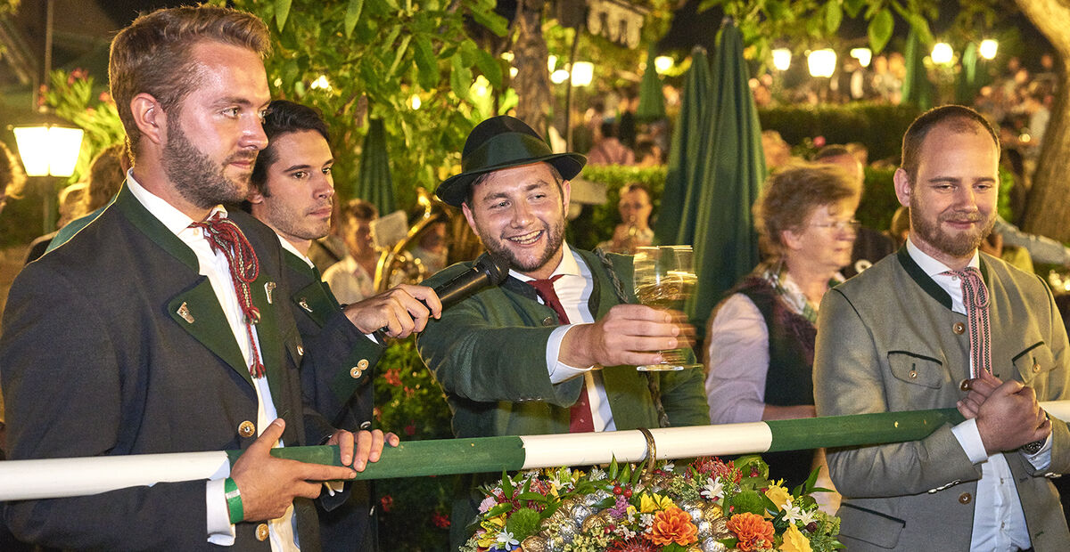 Vier Männer in traditioneller bayerischer Kleidung, einer hält ein Bierglas und lächelt bei einer Veranstaltung im Freien.