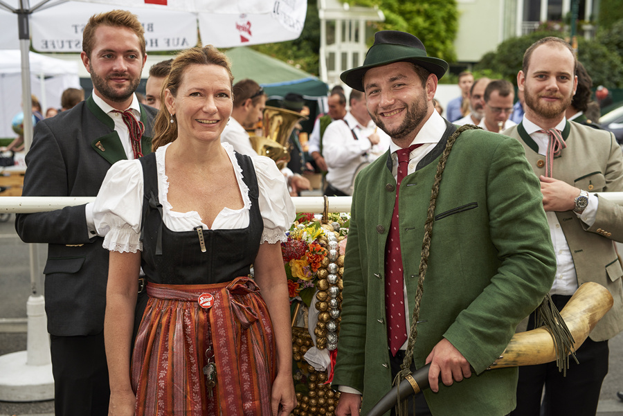 Vier Personen in traditioneller bayerischer Kleidung lächeln bei einem Fest im Freien, im Hintergrund sind weitere Teilnehmer zu sehen.