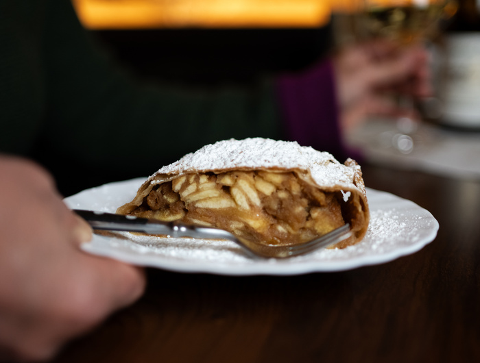 Eine Hand hält einen Teller mit einer Scheibe Apfelstrudel, die mit Puderzucker bestäubt ist, und einer Gabel an der Seite.