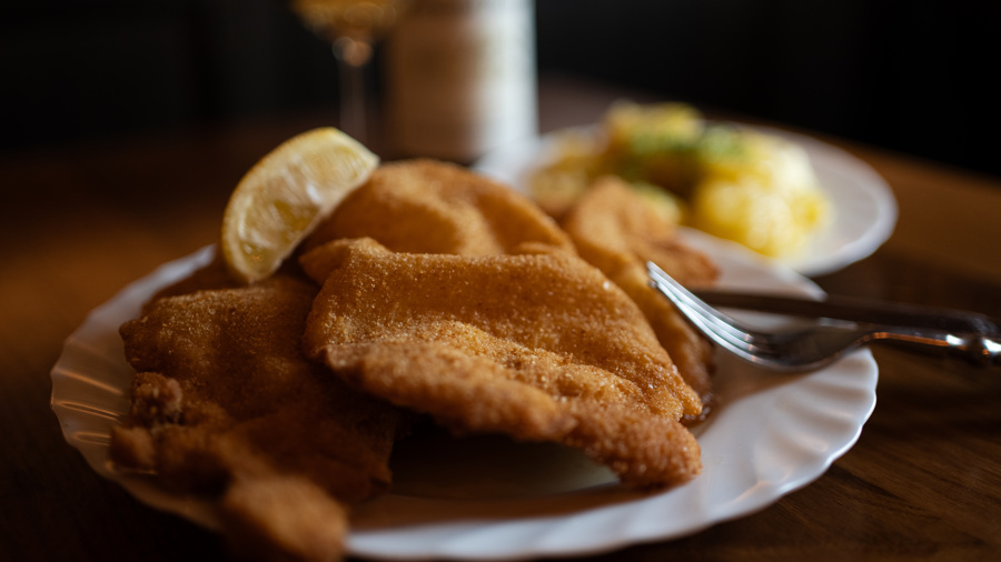 Paniertes Schnitzel mit einer Zitronenspalte auf einem weißen Teller, mit einer Gabel und einer Beilage im Hintergrund.
