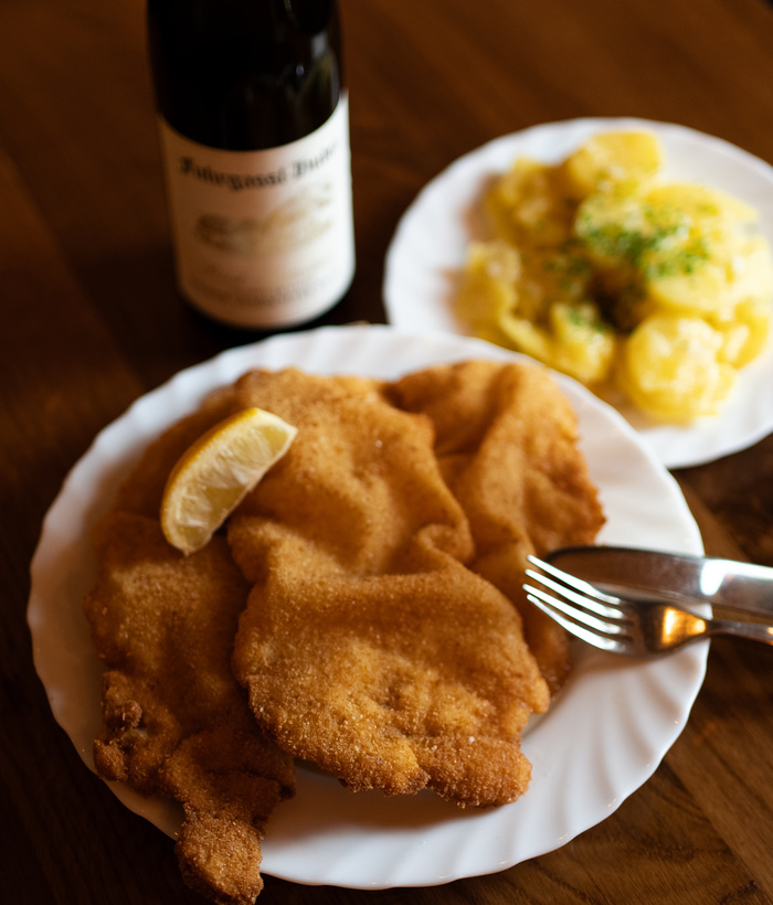 Knuspriges Schnitzel mit Zitrone, Kartoffelsalat und einer Flasche Wein auf einem Holztisch.