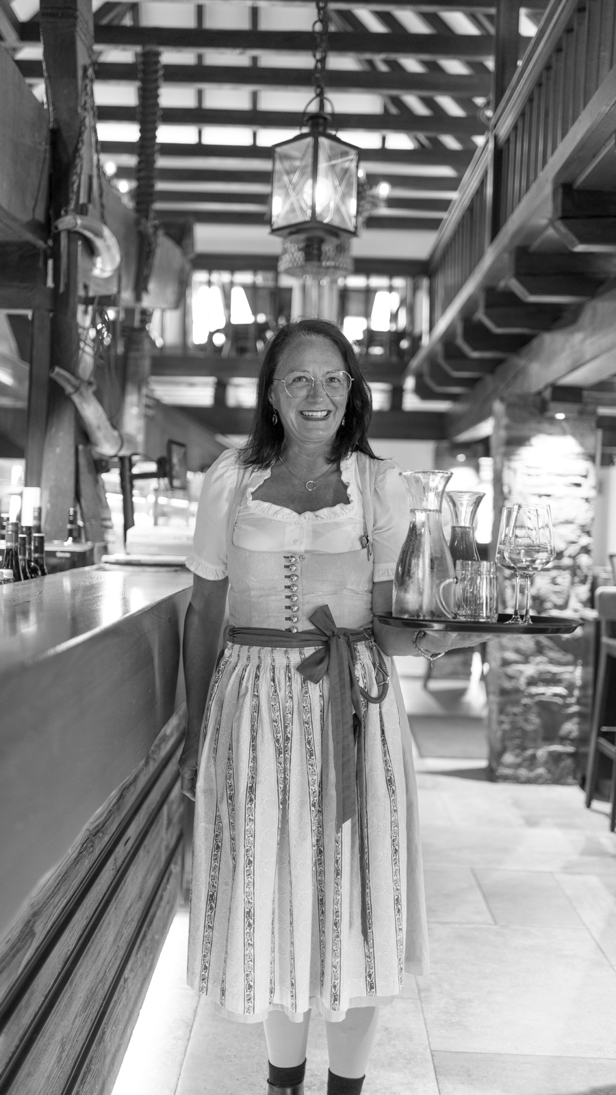 Eine lächelnde Frau in traditioneller Kleidung hält ein Tablett mit Gläsern und einer Karaffe in einem rustikalen Restaurant.