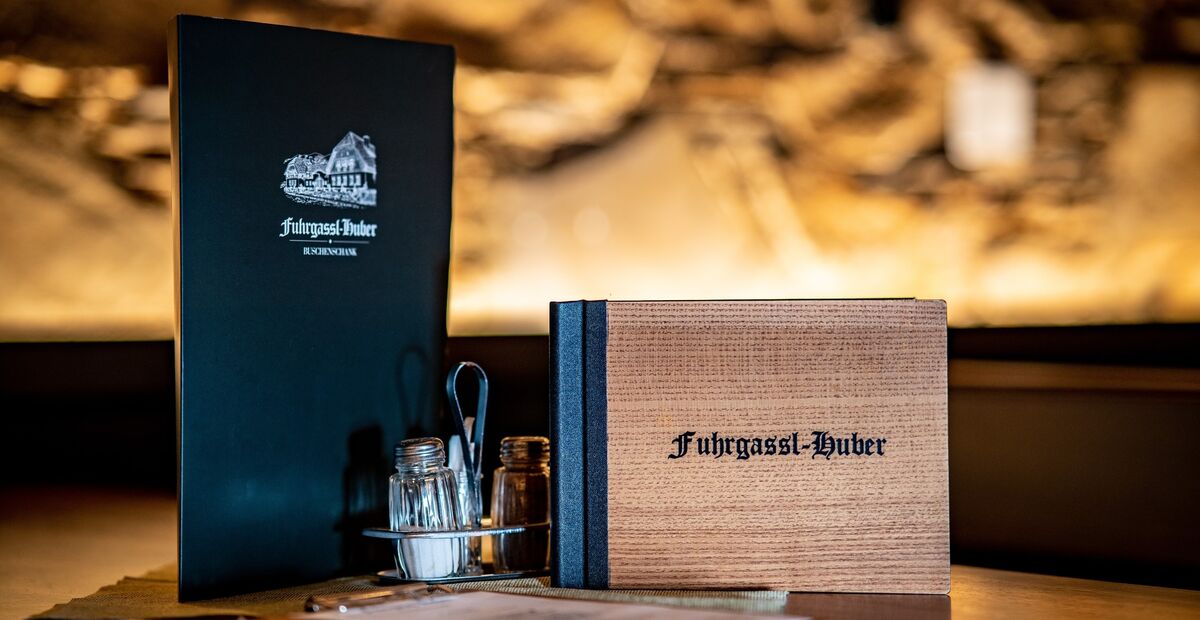 Hölzerne und schwarze Speisekarten mit der Aufschrift Fuxgassl-Huber mit Gewürzen auf einem Restauranttisch.