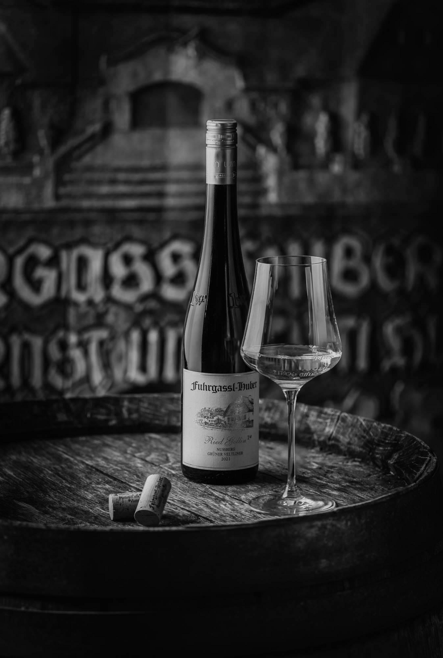 Eine Weinflasche, ein Glas und Korken stehen auf einem Holzfass mit verschwommener Schrift im Hintergrund.