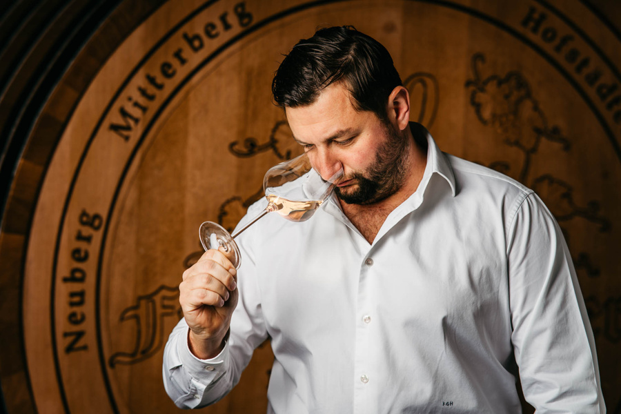 Mann in weißem Hemd riecht an einem Glas Wein und steht vor einem großen hölzernen Weinfass.