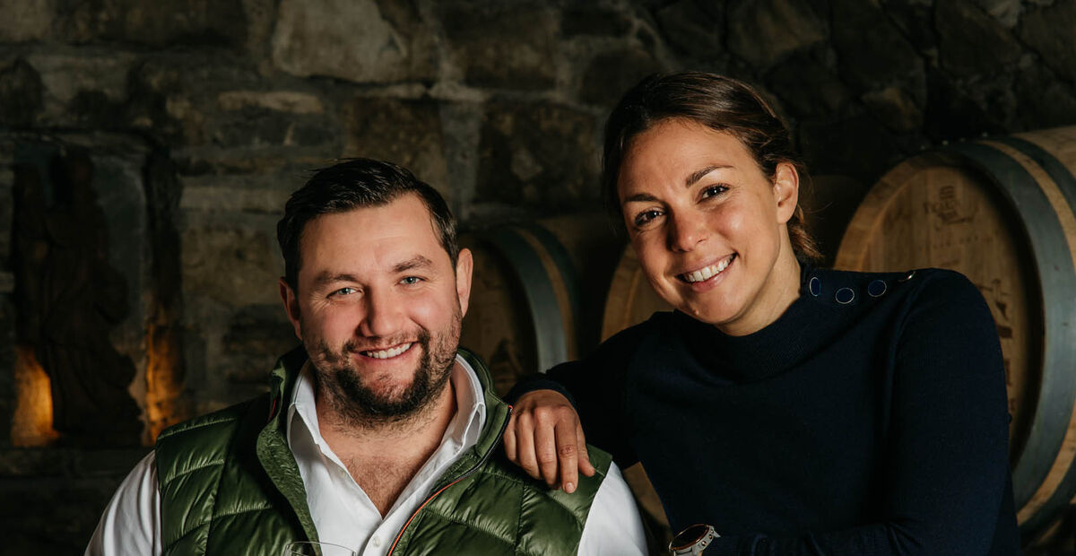 Ein Mann und eine Frau lächeln gemeinsam vor Weinfässern in einem rustikalen Keller.