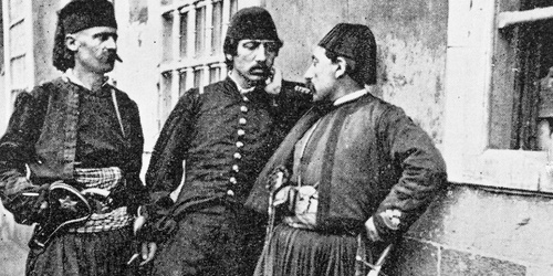 Drei Männer in traditionellen historischen Uniformen stehen vor einem Gebäude mit vergitterten Fenstern und unterhalten sich.