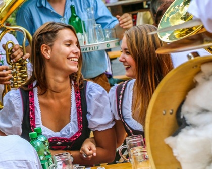 Zwei Frauen in bayerischen Trachten lachen bei einer festlichen Veranstaltung im Freien mit Getränken und Musikinstrumenten.